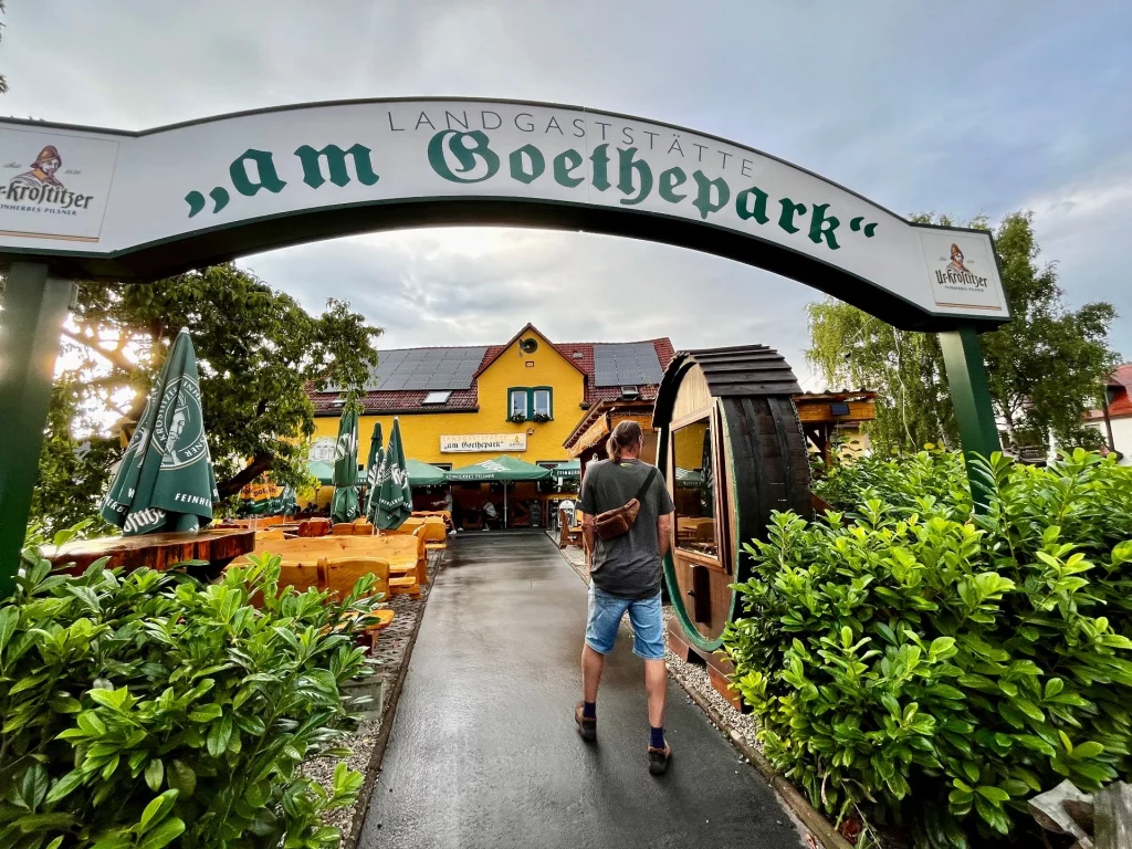 Eingangsbereich der Landgaststätte „am Goethepark“ in Jena. Ein großer weißer Torbogen mit der Aufschrift des Restaurantnamens rahmt den Blick auf einen gemütlichen Biergarten mit grünen Sonnenschirmen und ein gelbes Hauptgebäude mit Photovoltaikanlage ein.