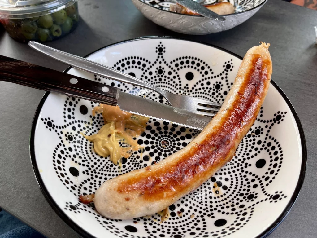 Eine fertig gebratene, goldbraune Bratwurst liegt auf einem weiß-schwarzen Teller mit Punktmuster. Daneben befindet sich ein Klecks Senf sowie Besteck (Messer und Gabel).