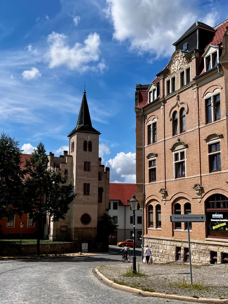 Blick in eine gepflasterte Straße in Naumburg. Links ein heller Kirchturm mit spitzem Dach, rechts ein prachtvolles Backsteingebäude mit verzierten Fenstern und einem Schild 'August-Reinstein-Haus'.