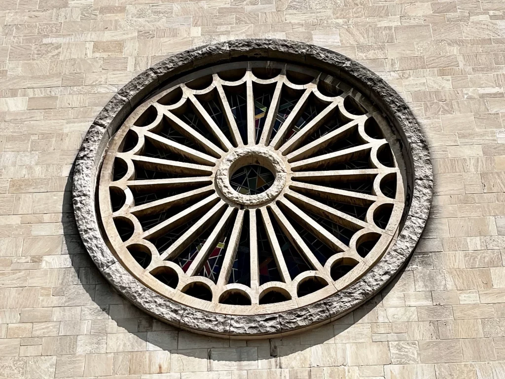 Nahaufnahme eines großen, steinernen Radfensters (Rosette) an einer hellen Fassade. Die Speichen des Fensters bilden ein symmetrisches Muster, dahinter ist buntes Glas erkennbar.