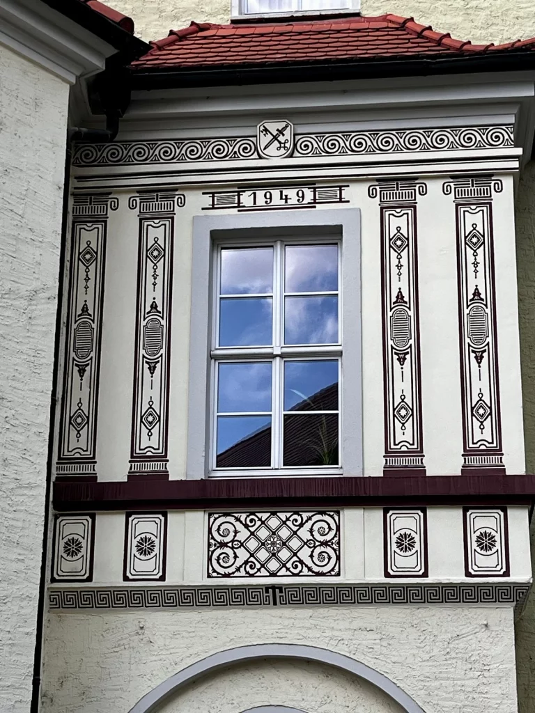 Detail einer hellen Hausfassade mit einem Fenster, das von dunklen, aufgemalten Säulen gerahmt ist. Über dem Fenster steht die Jahreszahl 1949 und ein kleines Wappen mit gekreuzten Schlüsseln und einem Schwert.