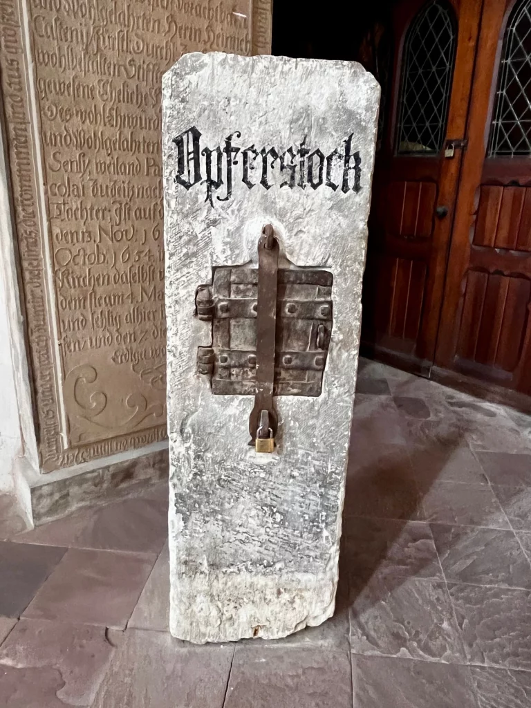 Ein massiver, aus hellem Stein gefertigter Opferstock mit der Aufschrift „Opferstock“ in schwarzer Frakturschrift. In der Mitte ist eine kleine, mit einem Vorhängeschloss gesicherte Eisentür eingelassen.