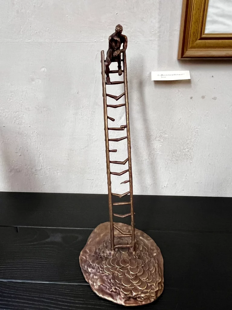 Schlanke Bronzeskulptur einer sitzenden Figur auf der Spitze einer sehr hohen, schmalen Leiter, montiert auf rundem Sockel.