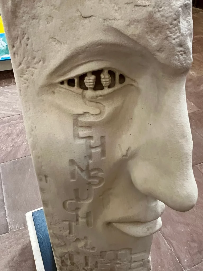 Steinerne Skulptur eines menschlichen Gesichts im Profil mit eingeschnittenem Auge und vertikal eingravierten Buchstaben auf der Wange.