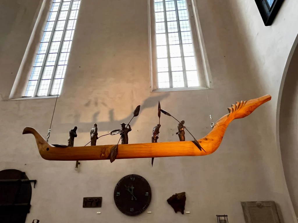 Hängende Holzskulptur eines stilisierten Boots mit mehreren kleinen Figuren und Rudern, ausgestellt in einem hohen Kirchenraum.