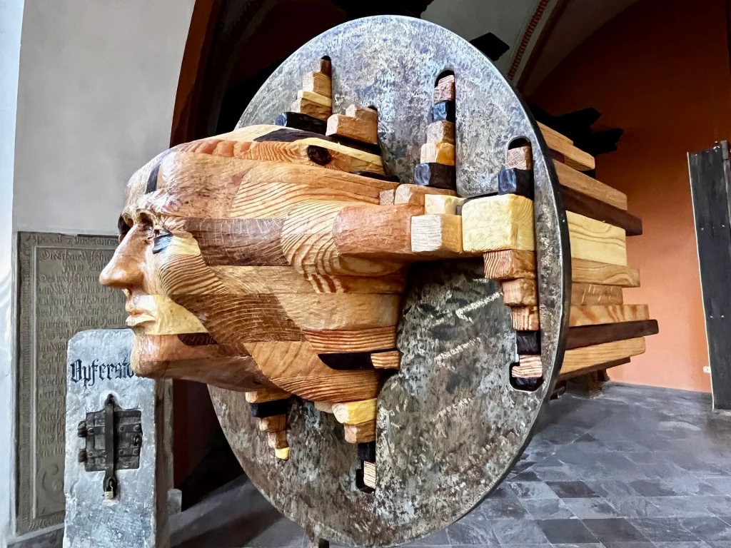Moderne Skulptur aus Holz und Stein in Form eines menschlichen Kopfes, zusammengesetzt aus Holzblöcken und von einer runden Steinscheibe durchzogen.