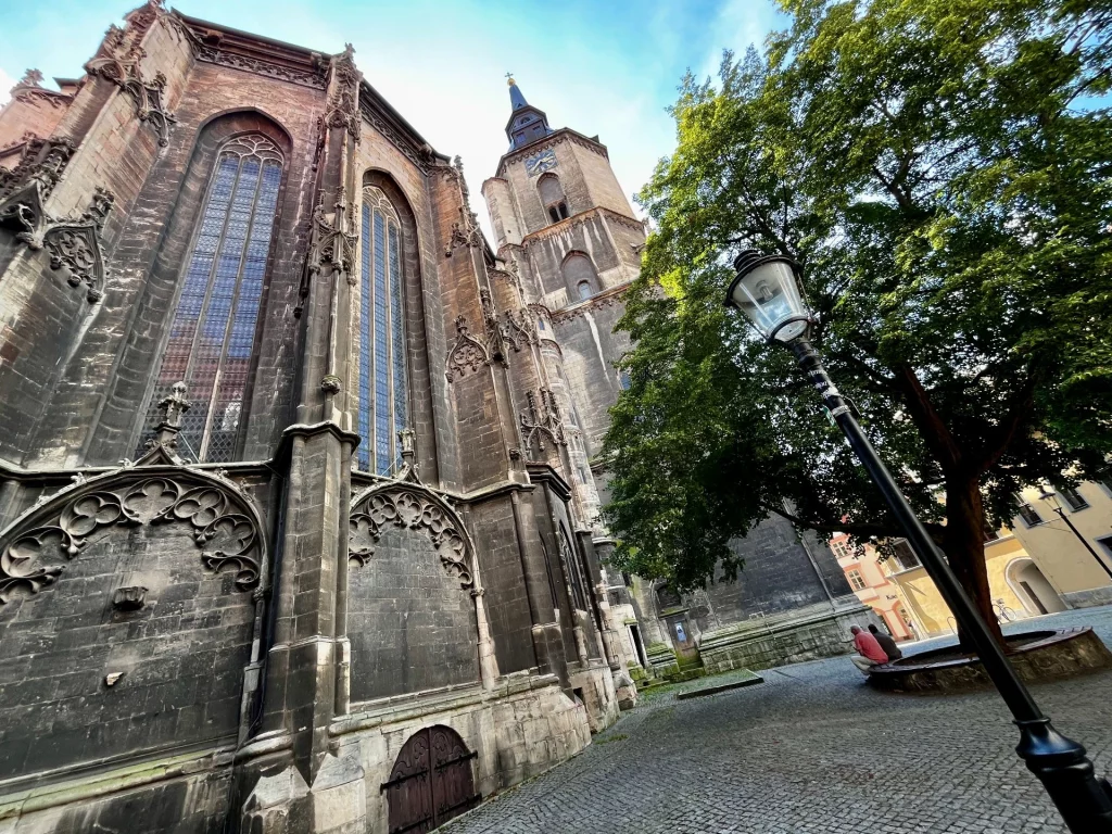 Eine beeindruckende Außenansicht der Stadtkirche St. Wenzel in Naumburg. Zu sehen ist eine massive, mittelalterliche Steinmauer mit vergitterten Fenstern und einer kleinen Holztür im Vordergrund. Dahinter ragt der markante Kirchturm mit seiner barocken Haube in den blauen Sommerhimmel.