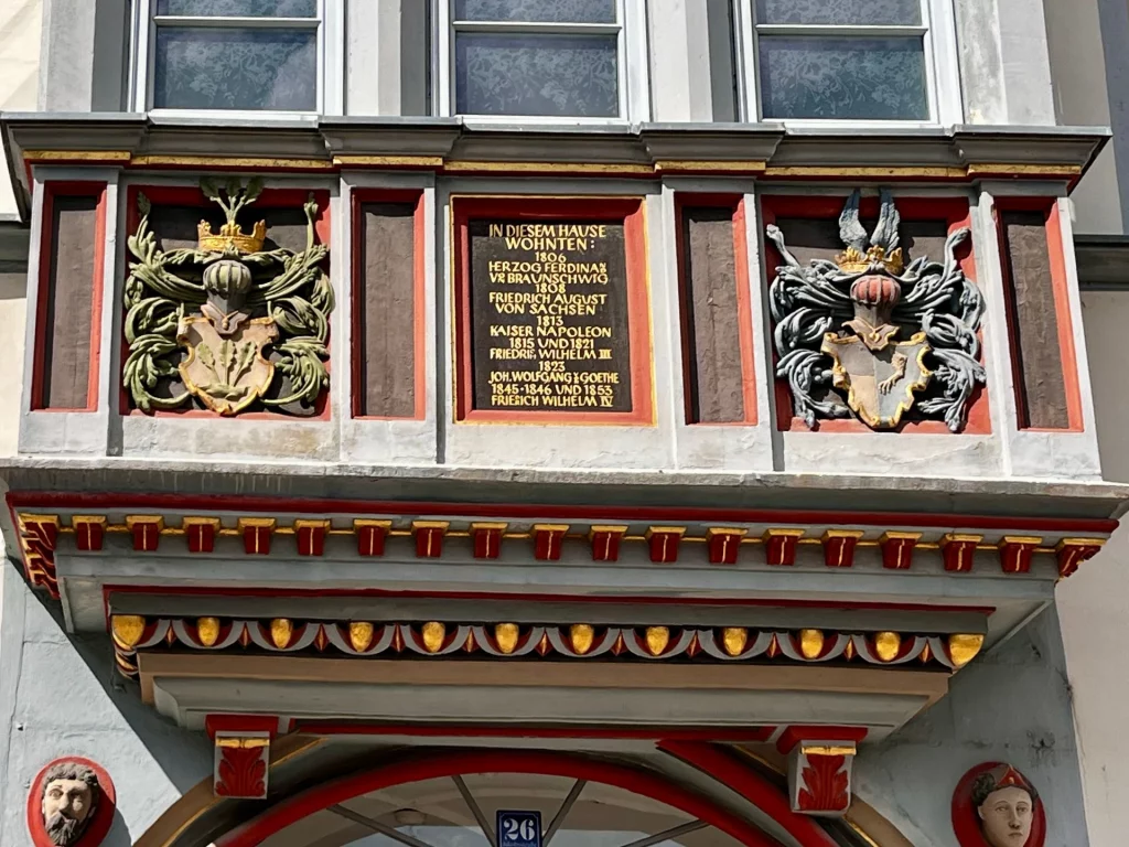 Detailansicht eines prunkvollen Erkers mit zwei farbigen Wappen und einer zentralen schwarzen Tafel in Goldinschrift, die berühmte Bewohner des Hauses wie Napoleon, Goethe und Friedrich Wilhelm IV. auflistet.