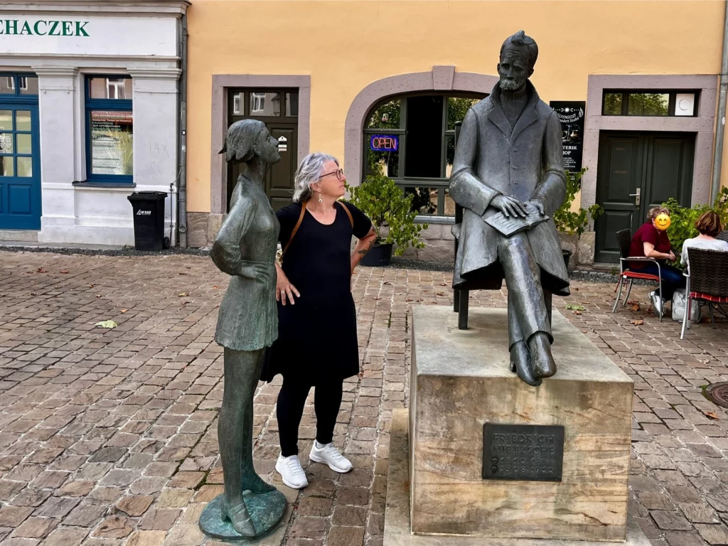 Auf einem kopfsteingepflasterten Platz steht Karin neben der lebensgroßen Bronzestatue des sitzenden Friedrich Nietzsche und blickt zu ihm auf.