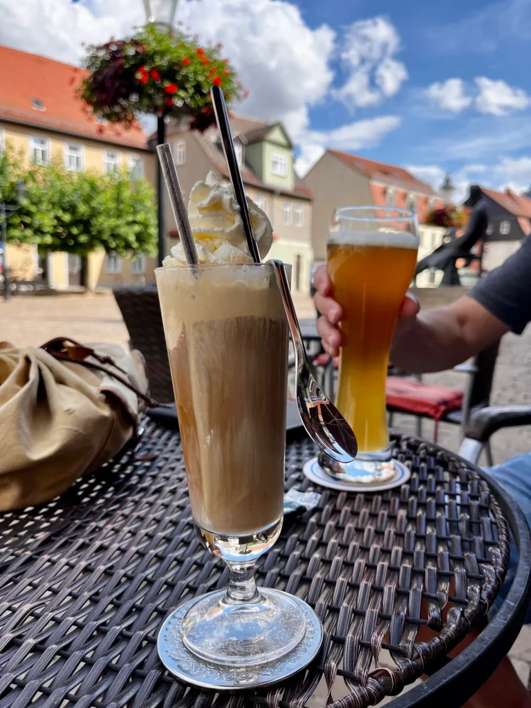 Café-Tisch im Freien mit einem hohen Glas Eiskaffee mit Sahne und einem Glas Bier, im Hintergrund bunte Häuser und Marktplatz.