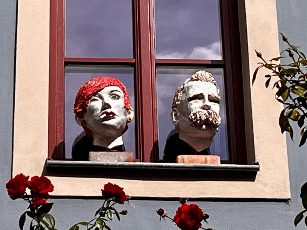 Zwei plastische Skulpturenköpfe auf einer Fensterbank, einer mit rotem Haar, der andere mit Bart, vor einem Fenster mit Blumen.