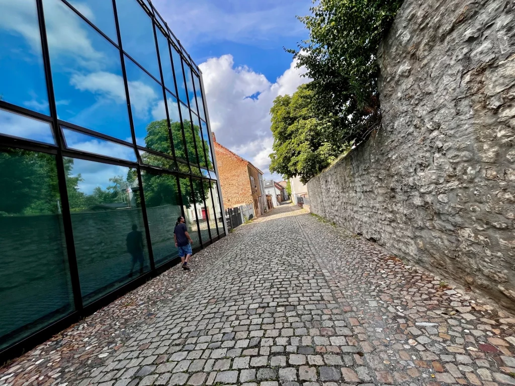 Kontrastreiche Gasse, in der sich auf der linken Seite die historische Architektur in einer modernen, blauen Glasfassade spiegelt, während rechts die raue, alte Stadtmauer aus Naturstein verläuft.