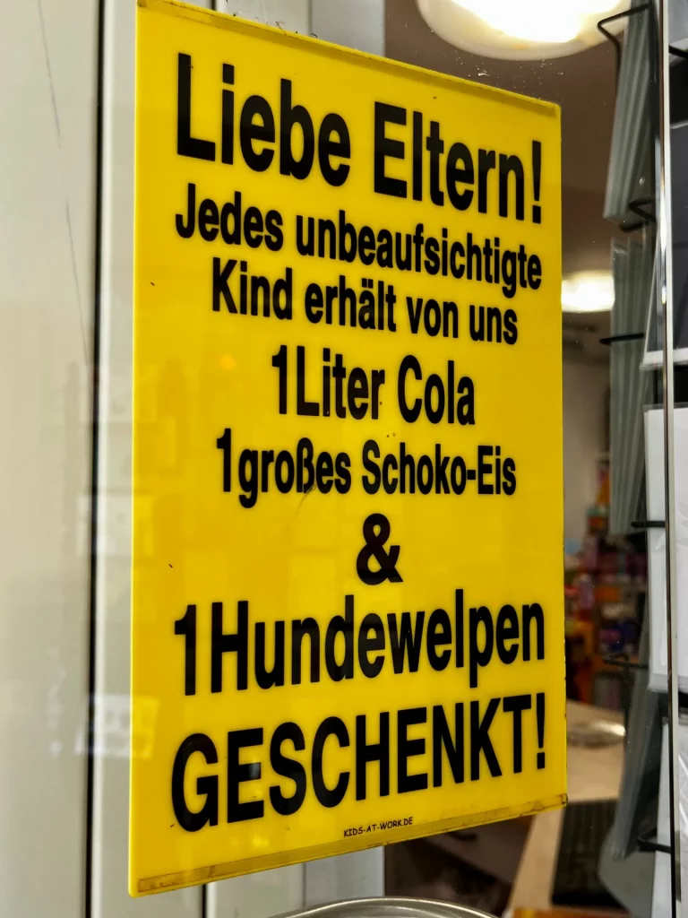 Gelbes Hinweisschild im Schaufenster mit humorvollem Text über unbeaufsichtigte Kinder, gedruckt in schwarzer Schrift.