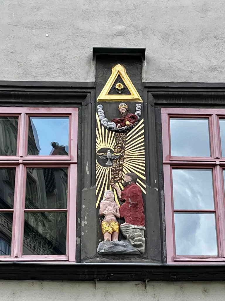 Relief an einer Hausfassade mit religiöser Szene, goldenen Strahlen und mehreren plastisch ausgearbeiteten Figuren.