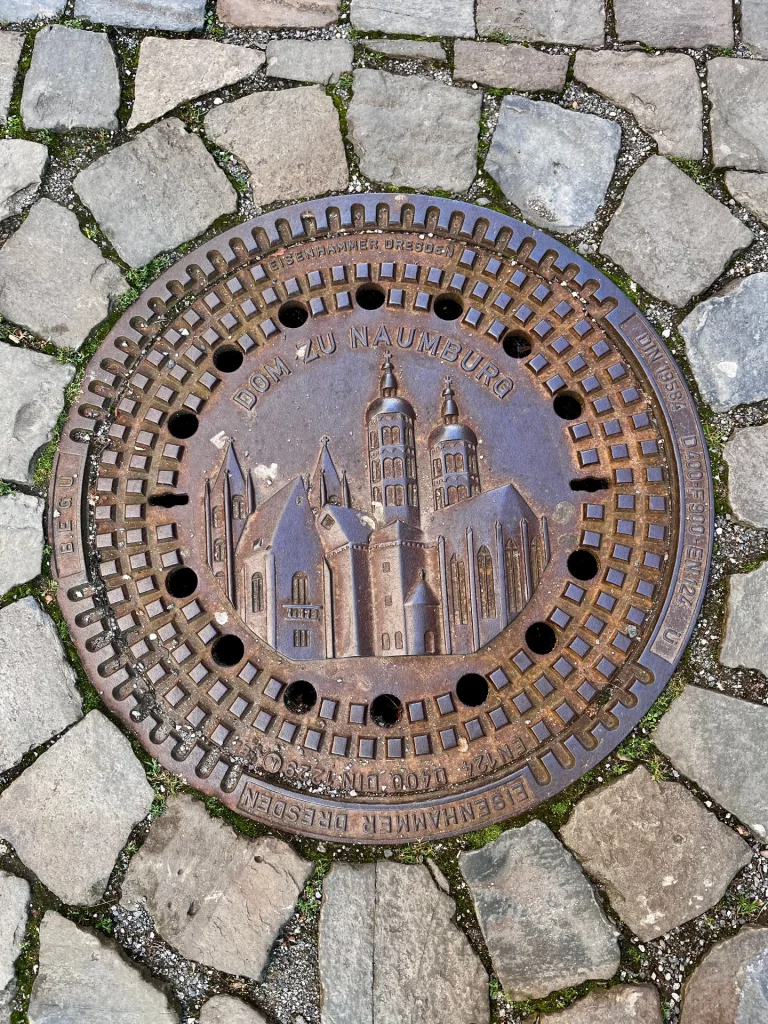 Runder Kanaldeckel im Kopfsteinpflaster mit Reliefdarstellung des Doms zu Naumburg und umlaufender Beschriftung.