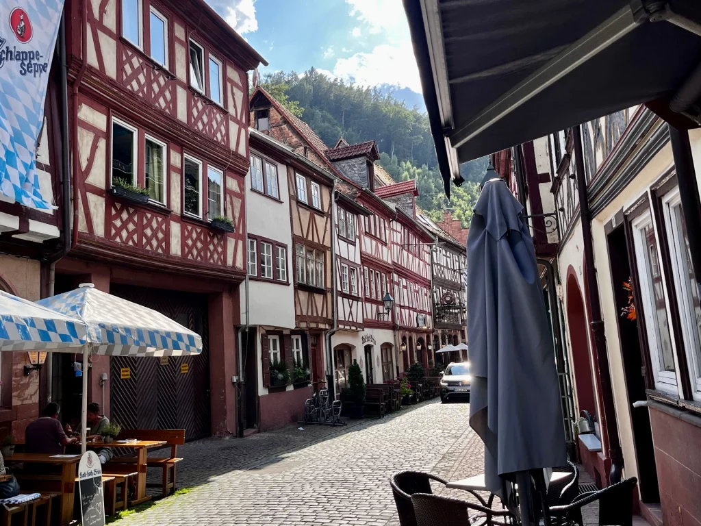 Perspektivische Ansicht einer gepflasterten Gasse mit dicht aneinandergereihten, historischen Fachwerkhäusern in Weiß- und Rottönen. Sonnenschirme der Außengastronomie säumen den Wegrand unter einem bewaldeten Hang.