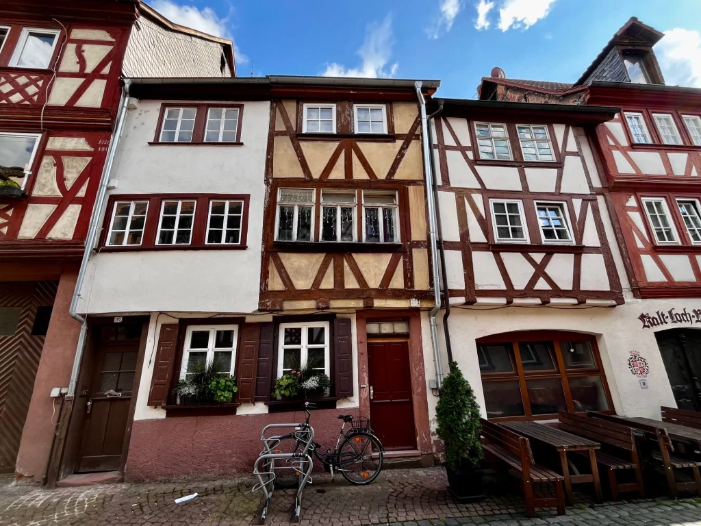Frontalansicht von drei schmalen, unterschiedlich gestalteten Fachwerkhäusern in Miltenberg. Die Fassaden zeigen klassische Holzständerbauweise mit Blumenkästen vor den Fenstern und abgestellten Fahrrädern davor.