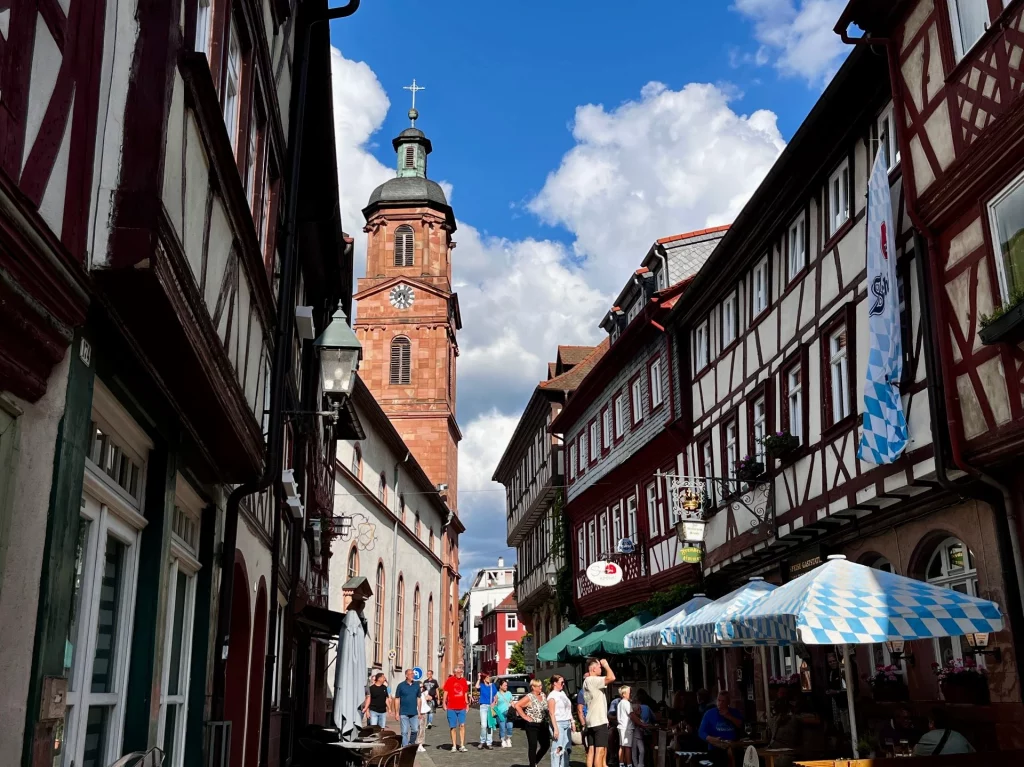 Eine schmale Gasse in der Miltenberger Altstadt mit hohen, prächtigen Fachwerkhäusern. Im Hintergrund ragt der rote Sandsteinturm der Stadtpfarrkirche St. Jakobus unter einem blau-weißen Wolkenhimmel empor.