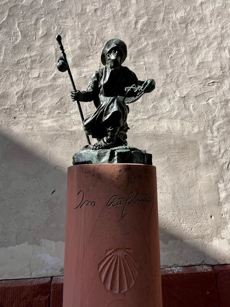 Eine Bronzestatue eines Wanderers oder Pilgers auf einer runden Sandsteinsäule mit der Aufschrift „Im Aufbruch“. Auf der Säule ist zudem eine Jakobsmuschel als Symbol des Jakobswegs eingraviert.