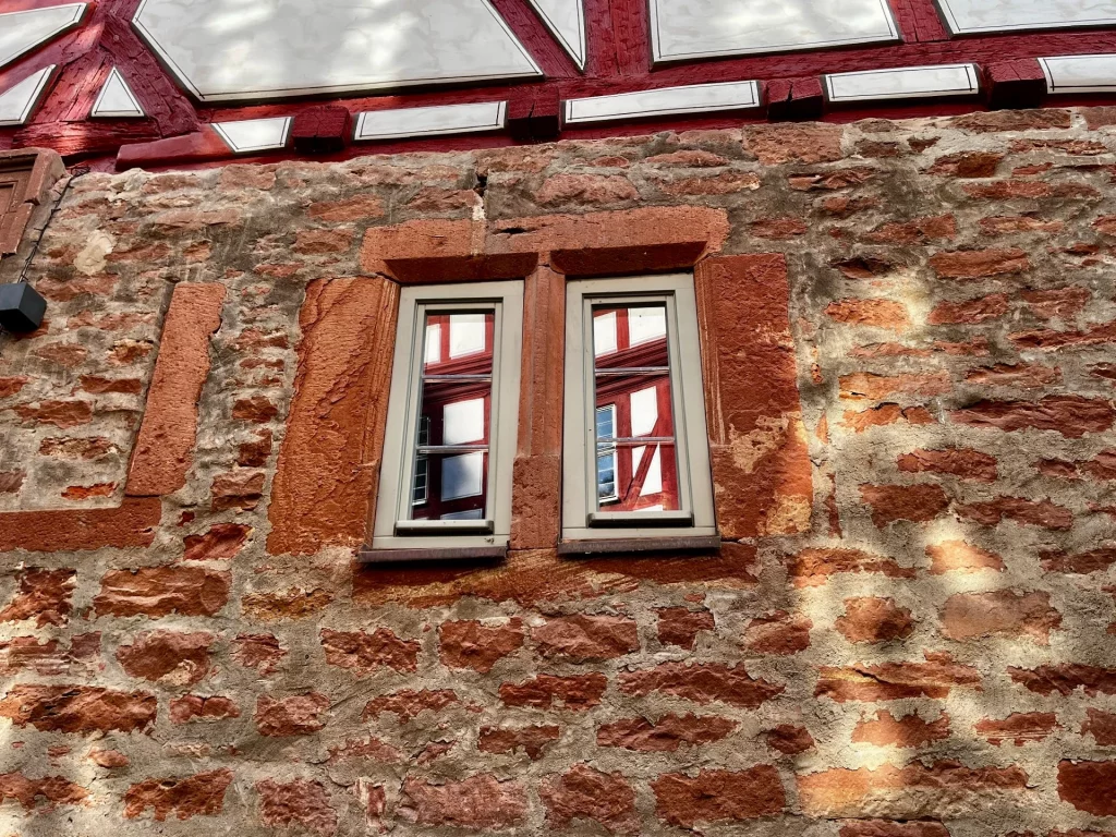 Zwei schmale, moderne Glasfenster, die in eine historische rote Sandsteinmauer eingelassen sind. In den Scheiben spiegelt sich das gegenüberliegende Fachwerk eines Hauses.