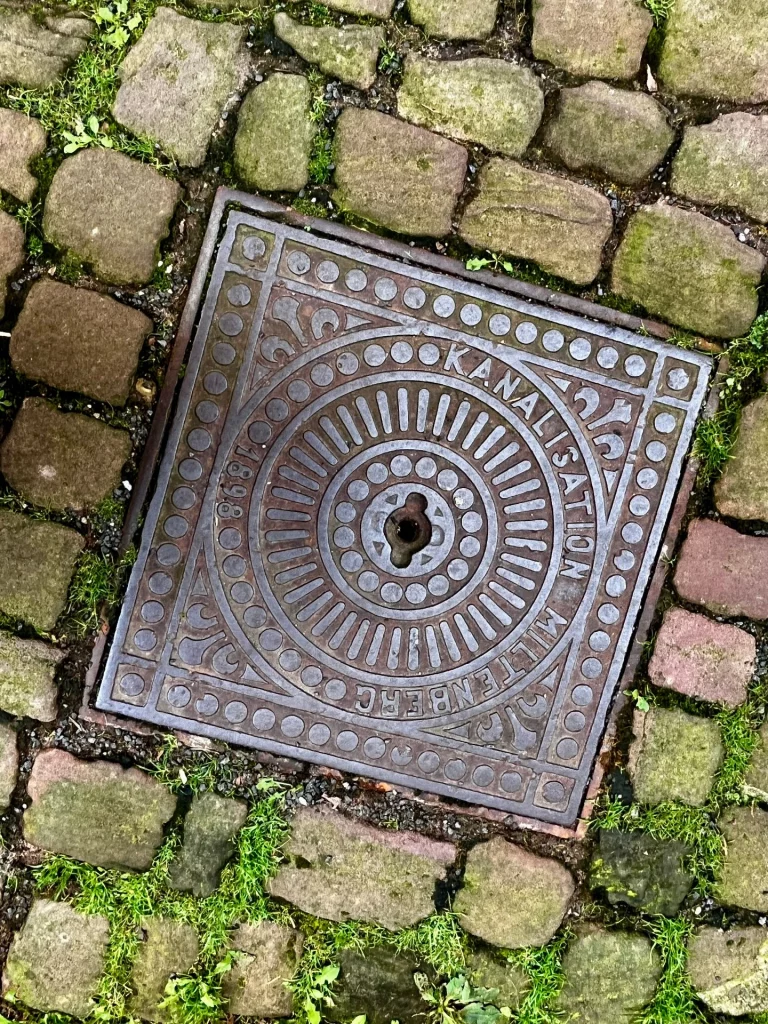 Ein quadratischer, gusseiserner Kanaldeckel im Kopfsteinpflaster mit der kreisförmigen Aufschrift „Kanalisation Miltenberg 1898“ und ornamentalen Verzierungen.