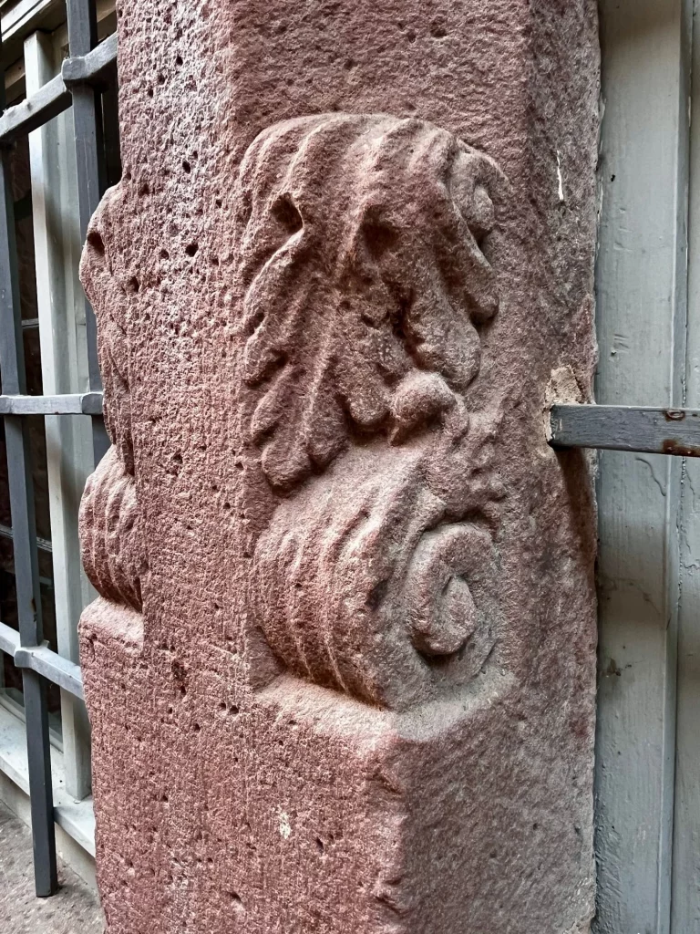 Nahaufnahme eines kunstvoll in roten Sandstein gehauenen Ornaments, das stilisierte Blätter und eine Schneckenform (Volute) an einer Hausecke oder Säule zeigt.