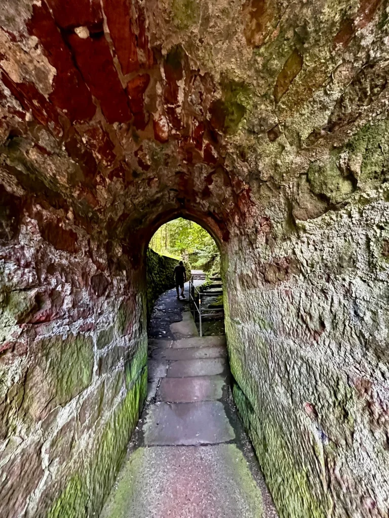 Blick durch einen engen, gewölbten Mauerdurchgang aus unregelmäßigem Stein auf einen weiterführenden Pfad, auf dem ein Wanderer im Schatten zu erkennen ist.