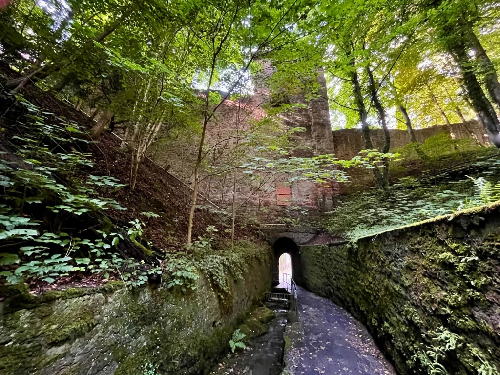 Blick durch einen engen, gewölbten Mauerdurchgang mit Turm aus unregelmäßigem Stein auf einen weiterführenden Pfad, auf dem ein Wanderer im Schatten zu erkennen ist.