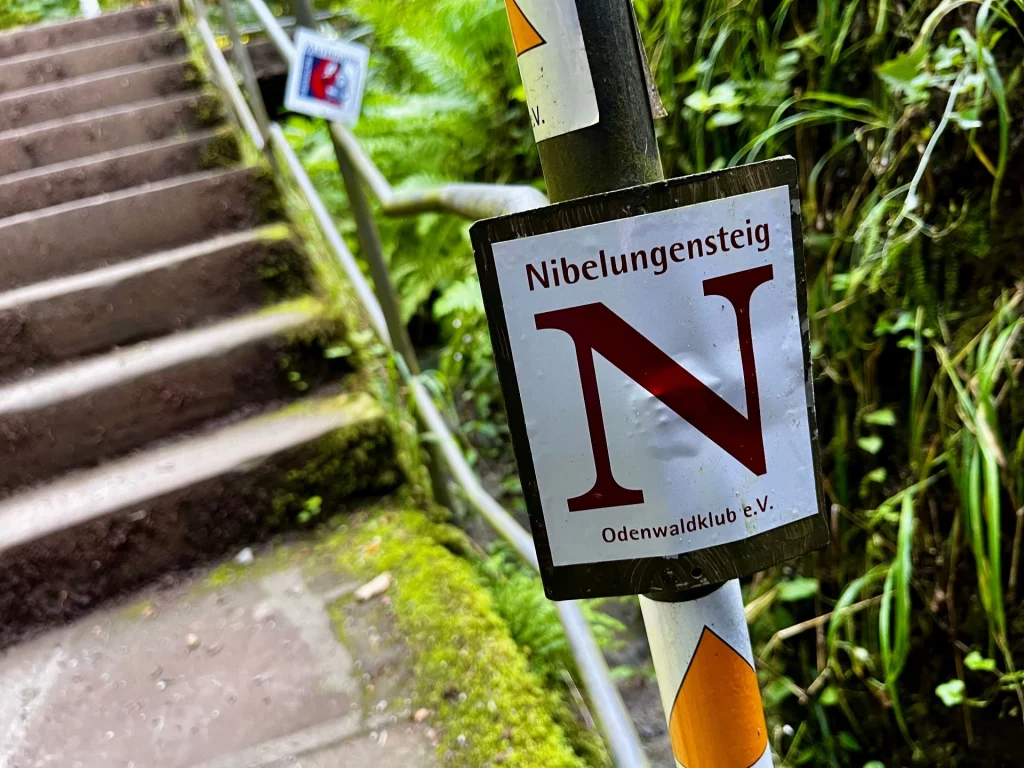 Nahaufnahme eines Wegweisers an einem Geländer mit dem roten Buchstaben „N“ auf weißem Grund für den Nibelungensteig, im Hintergrund sind steile Stufen zu sehen.