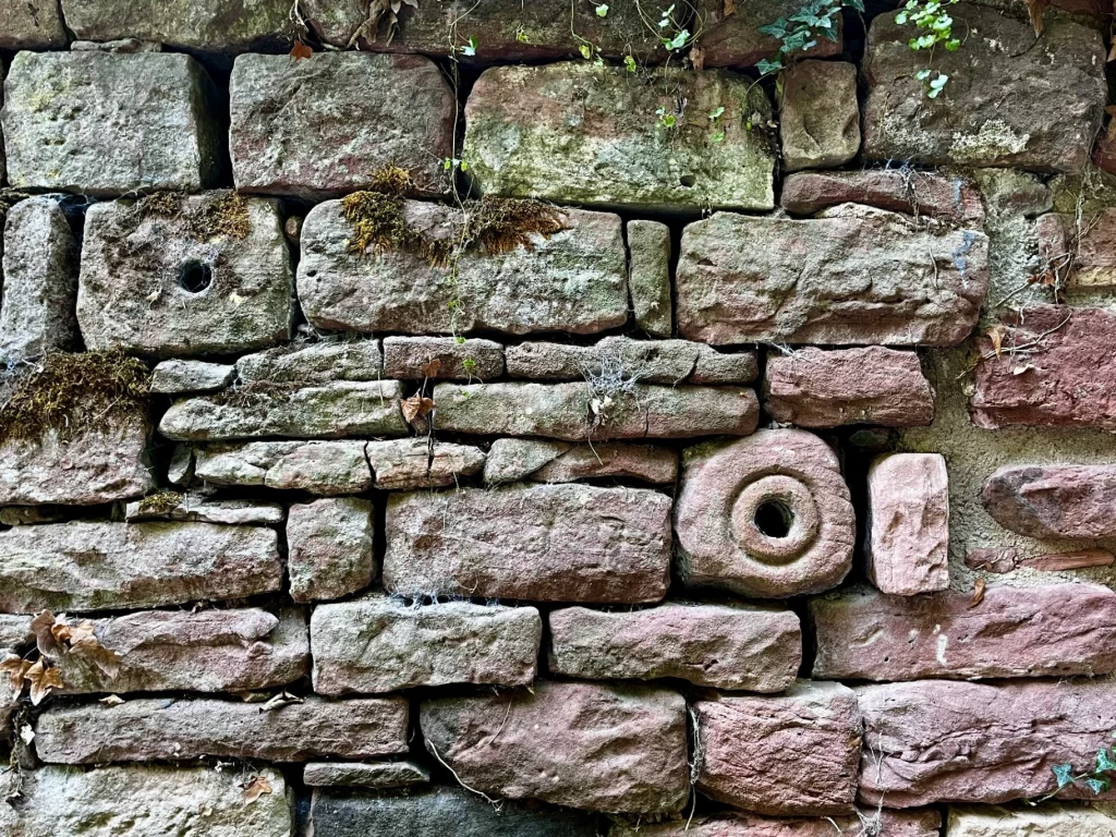 Detailansicht einer alten Mauer aus verschiedenfarbigen Sandsteinen, in die ein runder Stein mit einer kreisförmigen Öffnung – möglicherweise eine ehemalige Schießscharte oder ein Abfluss – eingelassen ist.
