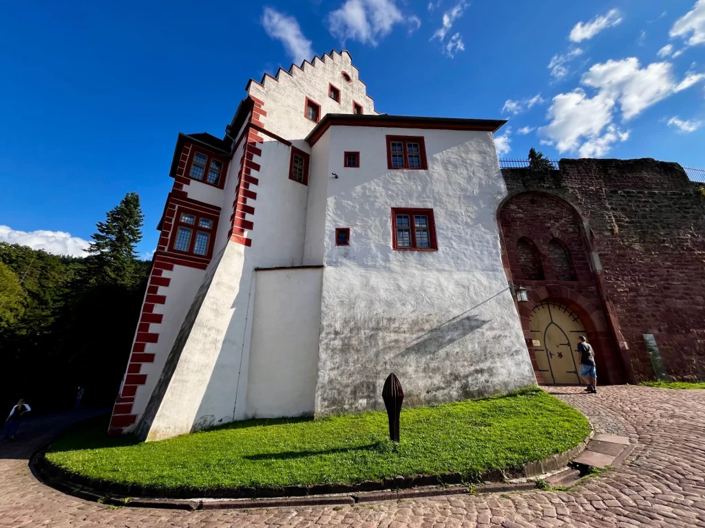 Die strahlend weiße Außenwand der Mildenburg mit markanten roten Eckquaderungen und Fensterrahmen unter einem tiefblauen Himmel. Ein gepflasterter Weg führt um das Gebäude herum zu einem massiven Sandstein-Eingangsportal, an dem ein Besucher steht.