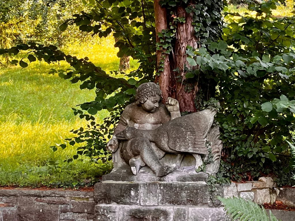 Verwitterte Sandsteinfigur vor Bäumen und Grünfläche. 