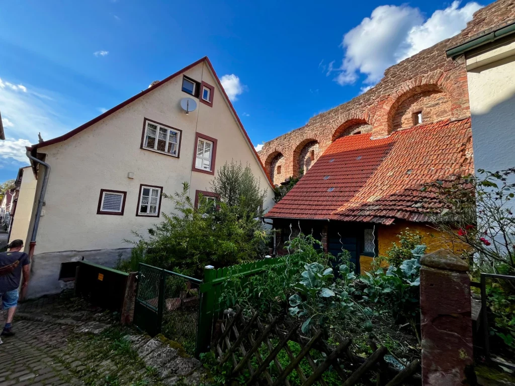 Altstadthäuser mit roten Ziegeldächern vor einer historischen Stadtmauer, umgeben von einem kleinen Garten.