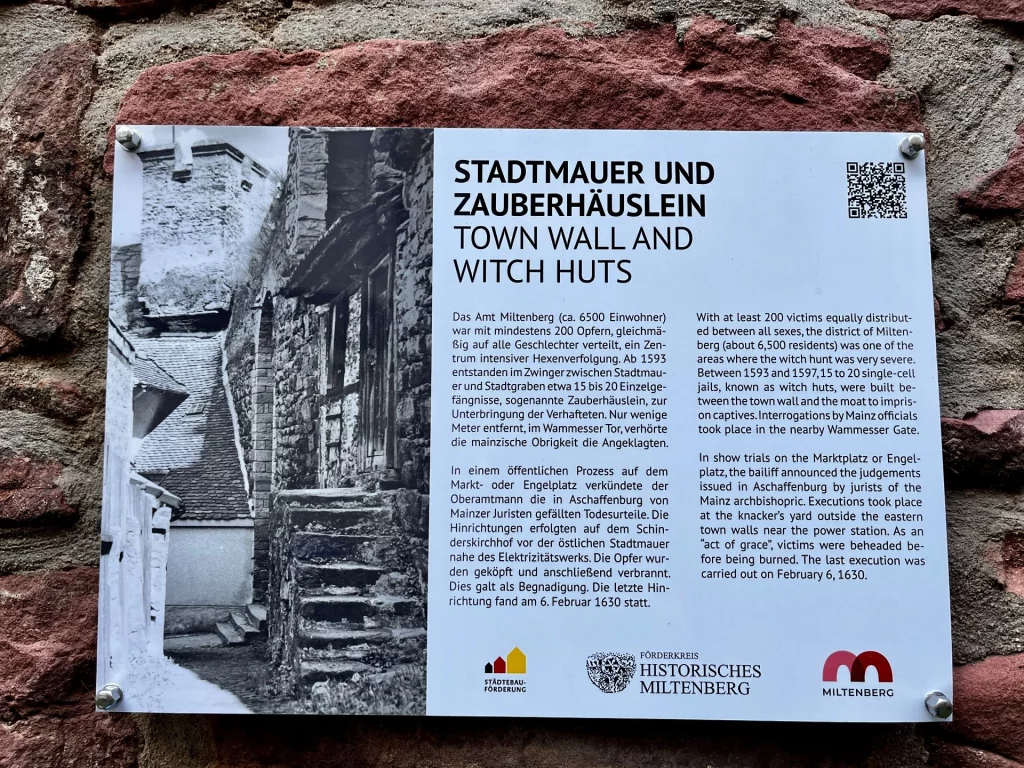 Eine Informationstafel mit dem Titel „Stadtmauer und Zauberhäuslein“ ist an einer rötlichen Sandsteinmauer befestigt. Die Tafel zeigt ein historisches Schwarz-Weiß-Foto von schmalen Gebäuden an einer Mauer und enthält deutschen sowie englischen Text über die Geschichte der Hexenverfolgung in Miltenberg.