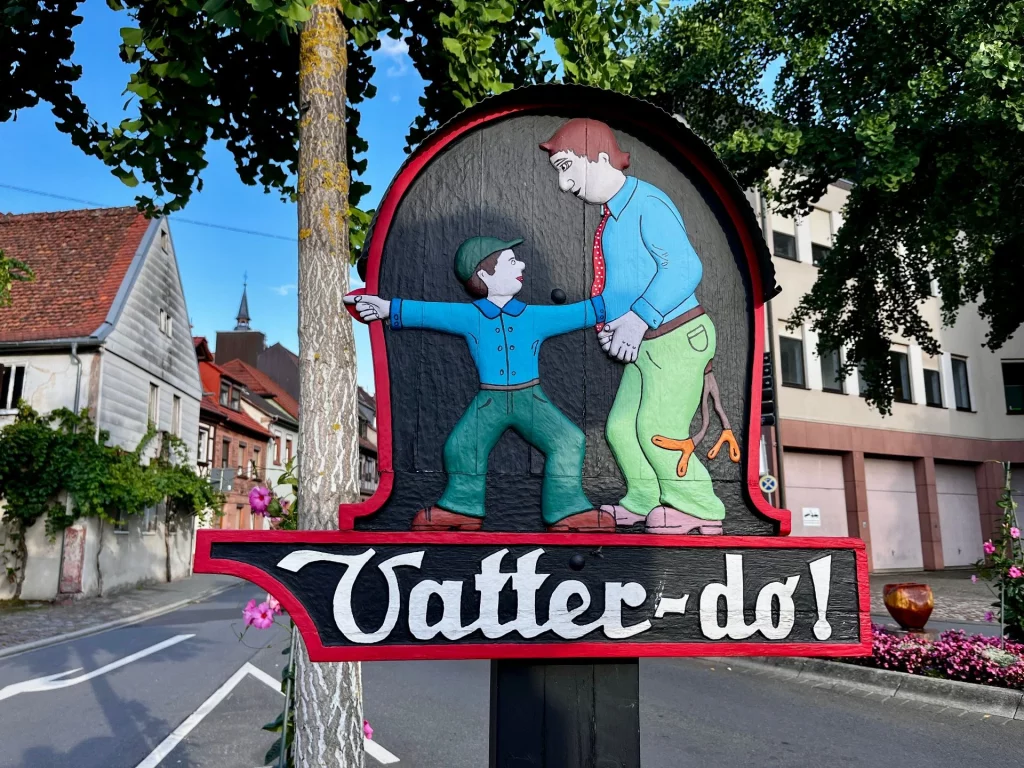 Bunt bemaltes Holzschild mit zwei Figuren und der Aufschrift „Vatter-do!“
