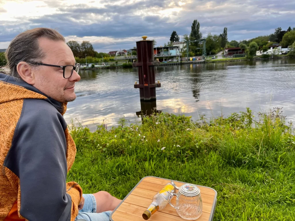 Eine Person sitzt am Flussufer auf einer Wiese und genießt die Ruhe. Auf einem kleinen Campingtisch stehen ein Glas und eine leere Flasche, im Hintergrund spiegelt sich der wolkige Himmel im Wasser des Mains.
