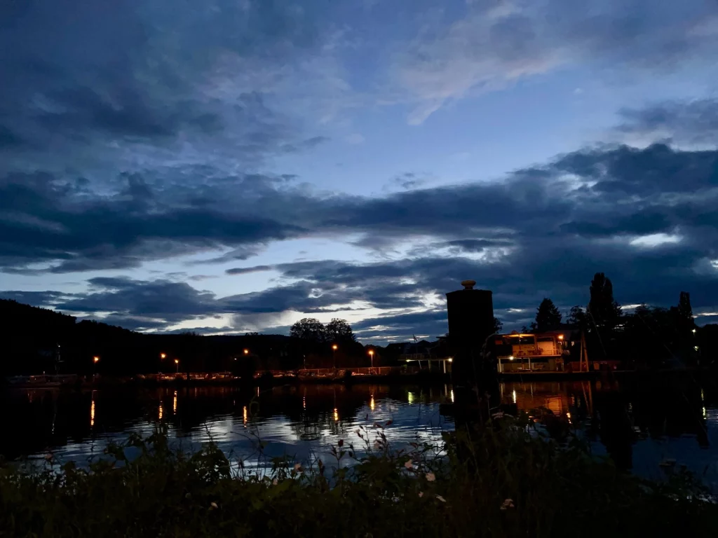 Abendstimmung am Fluss mit dunklen Wolken, beleuchteten Uferlichtern und deren Spiegelungen im ruhigen Wasser.