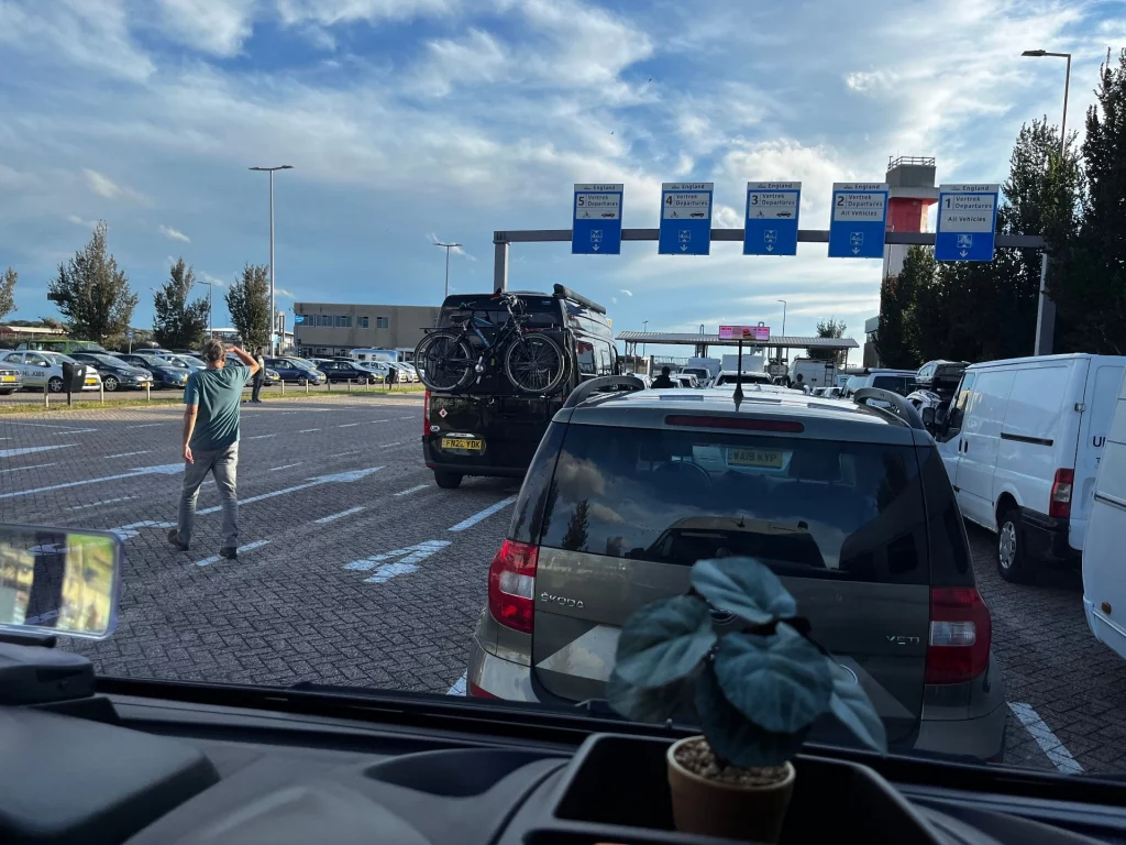 Blick aus dem Cockpit eines Fahrzeugs auf eine Warteschlange im Hafen von Hoek van Holland; im Hintergrund Hinweisschilder für die Abfahrt nach England.