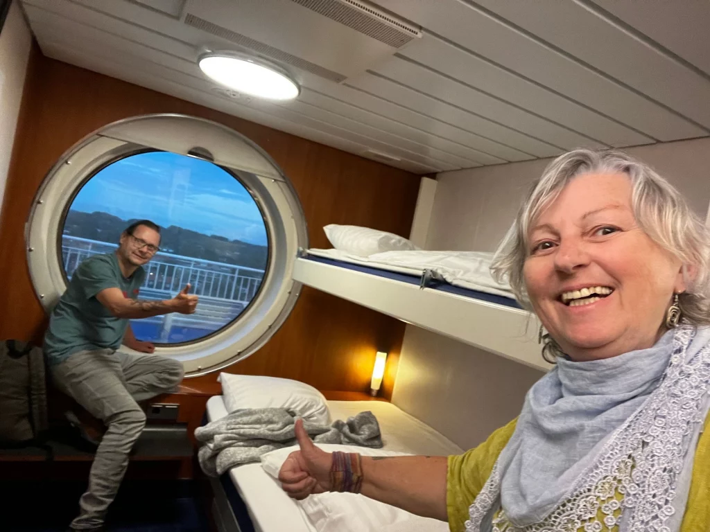 Ein gut gelauntes Paar in ihrer Schiffskabine; im Hintergrund ein großes, rundes Bullauge mit Blick auf das Meer und ein Doppelstockbett.