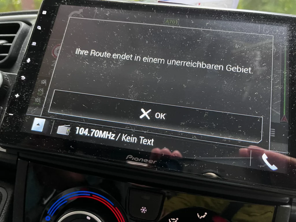 Nahaufnahme eines Pioneer-Navigationssystems im Cockpit eines Fahrzeugs mit der Fehlermeldung: „Ihre Route endet in einem unerreichbaren Gebiet.“ Im Hintergrund ist durch die Windschutzscheibe eine Straße im Grünen erkennbar.