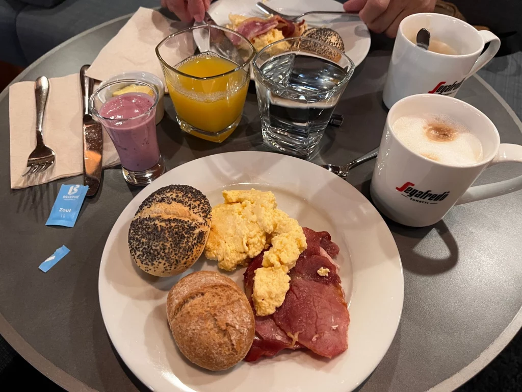 Draufsicht auf einen Frühstückstisch mit zwei Tellern: Rührei mit Speck und verschiedenen Brötchen. Dazu stehen Orangensaft, ein Glas Wasser, ein Beerensmoothie sowie zwei Tassen Kaffee der Marke Segafredo auf dem grauen Tisch.