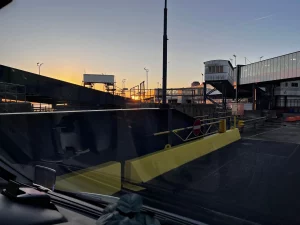 Blick aus der Windschutzscheibe eines Fahrzeugs auf das Hafengelände von Harwich bei Sonnenaufgang. Die goldene Sonne steht tief am Horizont hinter Brücken- und Terminalgebäuden.