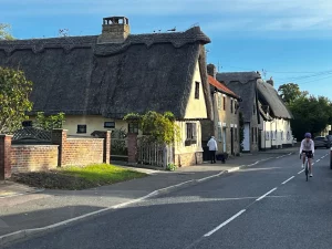 Eine Dorfstraße in Wilbraham mit historischen, hellgelben Häusern, die dicke, kunstvoll gedeckte Strohdächer (Reetdächer) tragen. Eine Radfahrerin und ein Fußgänger sind auf der sonnigen Straße unterwegs.
