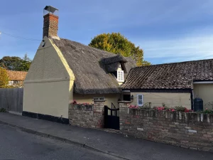 Nahaufnahme eines traditionellen Hauses mit Reetdach in Wilbraham. Eine niedrige Backsteinmauer mit rosa Blumen säumt das Grundstück unter einem strahlend blauen Himmel.