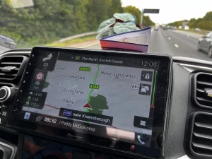 Das Pioneer-Navigationssystem im Fahrzeug zeigt eine Karte der A1(M) nahe Knaresborough. Auf dem Armaturenbrett liegt eine kleine Pflanze und eine gefaltete Broschüre; im Hintergrund ist die Autobahn bei Tageslicht erkennbar.