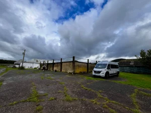 Der graue Kastenwagen (Kätter) steht geparkt auf dem asphaltierten Hof der Burnside Farm unter einem dramatischen, wolkenverhangenen schottischen Himmel.