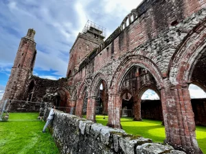 Mittelalterliche Architektur der Sweetheart Abbey bei Dumfries: Blick durch die Kirchenruine auf das Hauptschiff und den Vierungsturm unter bewölktem Himmel.