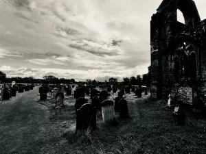 Schwarzweißaufnahme des Friedhofs der Sweetheart Abbey mit schiefen Grabsteinen im Vordergrund und den teilweise erhaltenen Mauern der Klosterruine unter einem dramatisch bewölkten Himmel.
