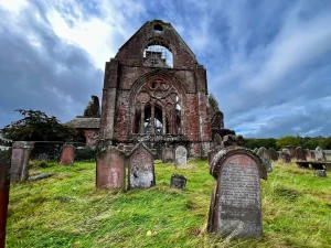 Farbaufnahme der Sweetheart Abbey mit der markanten gotischen Westfassade aus rotem Sandstein, umgeben von historischen Grabsteinen auf einer grünen Wiese unter blauem Himmel mit Wolken. 