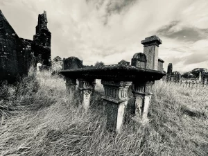 Ein seltener ‚Table Tomb‘ (Tischgrab) auf hohen, verzierten Steinbeinen, umgeben von hohem Gras auf dem historischen Friedhof; im Hintergrund ragen die dunklen Mauern der Abtei auf.
